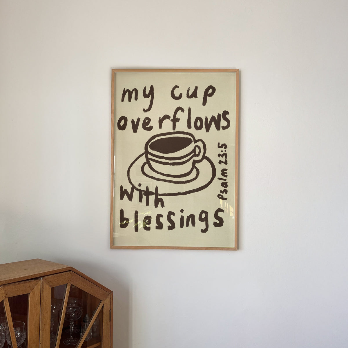 A framed art print with the text 'my cup overflows with blessings Psalm 34:5' and a simple coffee cup illustration, hanging on a white wall above a wooden cabinet.